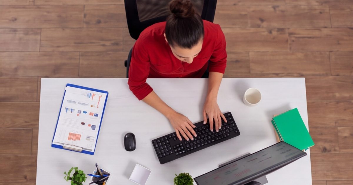 An executive assistant in a red top working on her laptop with a clipboard to her right