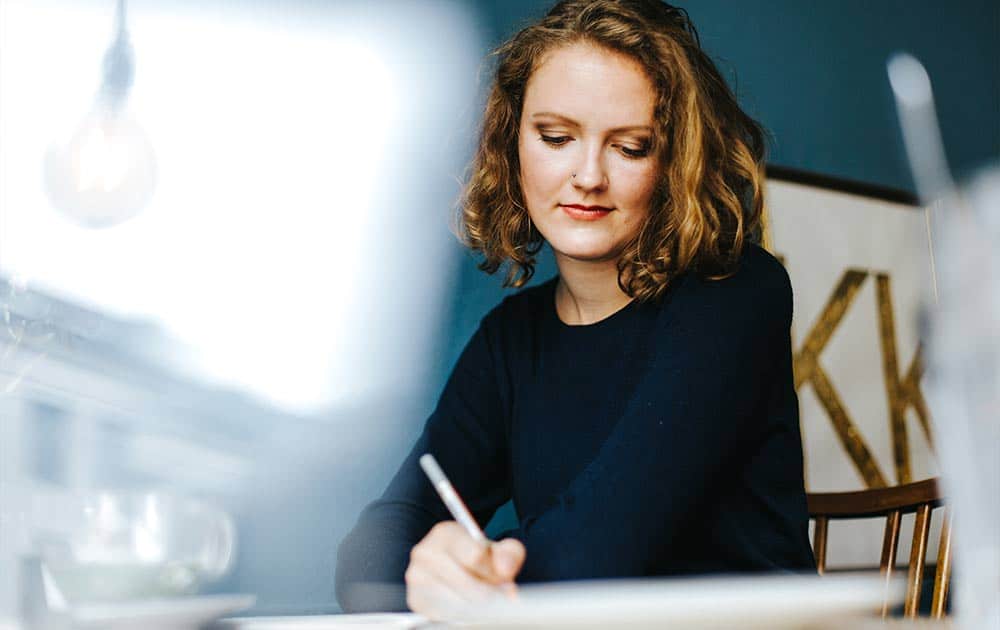 A happy professional woman sits at her desk, thoughtfully writing her career and personal goals in a planner.