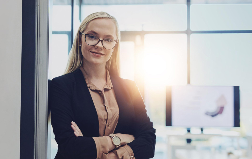 A composed and confident executive assistant standing in a modern corporate office, representing the interpersonal skills and discretion required for the role.